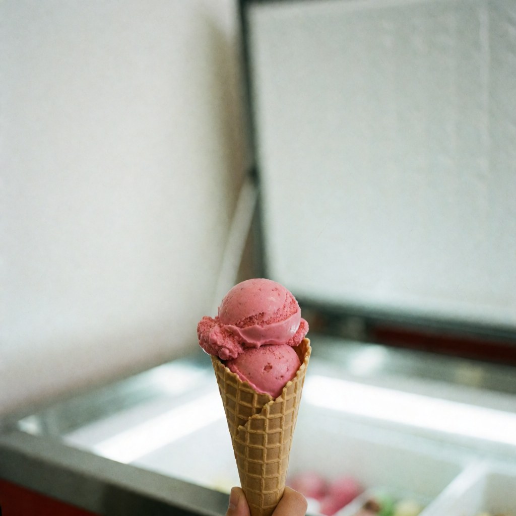 A pink strawberry ice cream cone held in front of a bright ice cream shop display case with a white background.