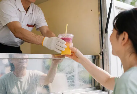 A worker wearing gloves hands a colorful shake to a customer from a beige mobile vendor truck during the day, with reflections visible on the window.