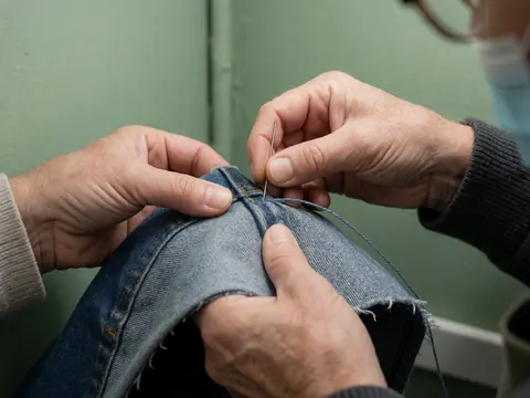 Adult hand sewing denim pants with thick thread and needle against a green wall, showing visible fabric texture and professional lighting