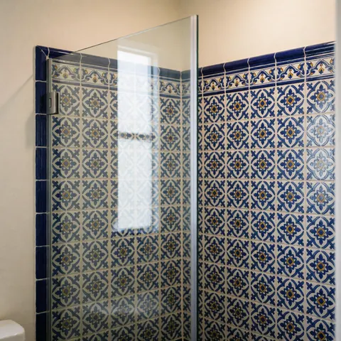 Transparent frameless glass shower door installed against blue and white Talavera tiles with decorative patterns in a modern bathroom.