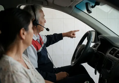 Instructor wearing a headset teaching inside a car with white tiled walls while a client listens attentively during a realistic driving service scene