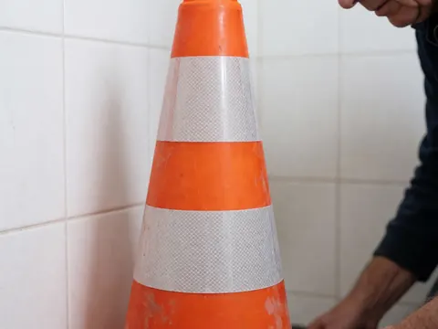 Close-up of an orange traffic cone with reflective bands being adjusted by a worker's hand against a white tiled wall