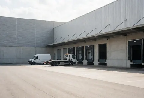 White tow truck and pickup truck parked in front of empty industrial loading docks with natural light on concrete pavement