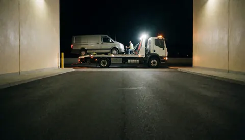 White flatbed tow truck carrying a van on a paved surface at night with bright lights and beige walls nearby