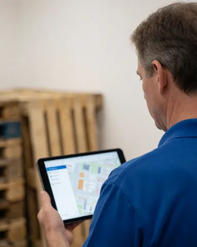 A man in a blue shirt reviews a map on a tablet in a warehouse with stacked wooden pallets in the background.