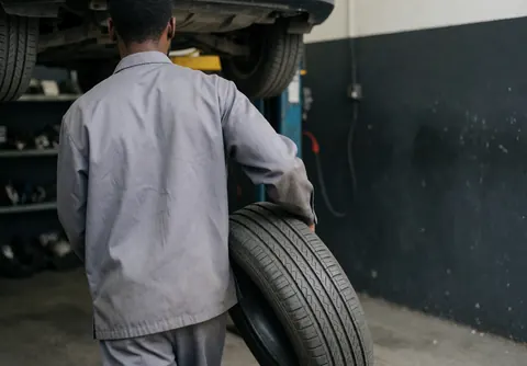 Mechanic carrying a tire toward a car on a lift in a repair shop with dark walls and shelves of parts in the background