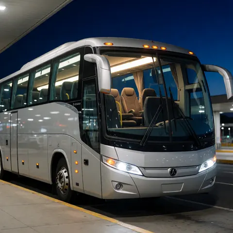 White modern tour bus parked at a night terminal with interior lights on and reflections on the windows.