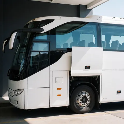 White tour bus parked at terminal with gray walls, tinted windows, and visible wheels under sunlight.