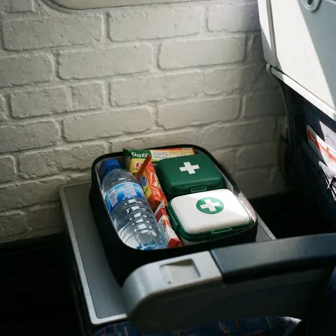 Travel kit containing water, snacks, and first aid supplies arranged on a bus seat against a white brick wall with natural lighting