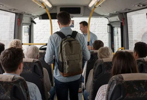 A tourist carrying a backpack steps onto a tour bus while a guide assists. Visible seats and white brick walls create a welcoming atmosphere inside the vehicle.