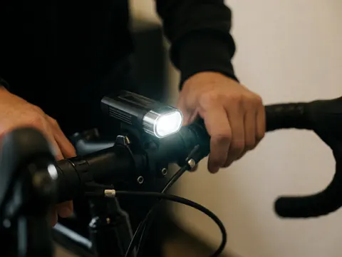 Close-up of a bright LED light on a bicycle handlebar with hands adjusting the cable against a dark background and warm wall.