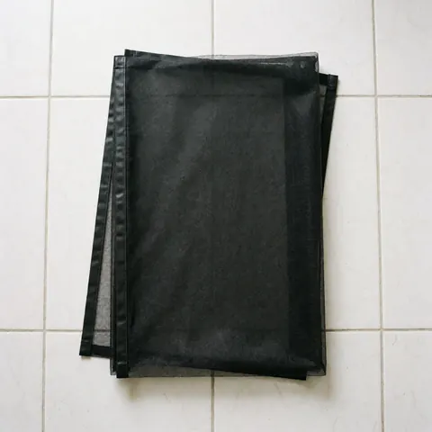 Folded black magnetic window screen with magnetic strips lying on white tiled floor ready for installation