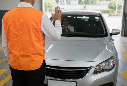 A parking worker in an orange vest guides a silver car with hand gestures. Yellow arrows mark the concrete floor near a garage door letting in natural light.