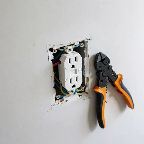 Newly installed white electrical outlet on a white wall with visible multicolored wires and black and orange pliers nearby