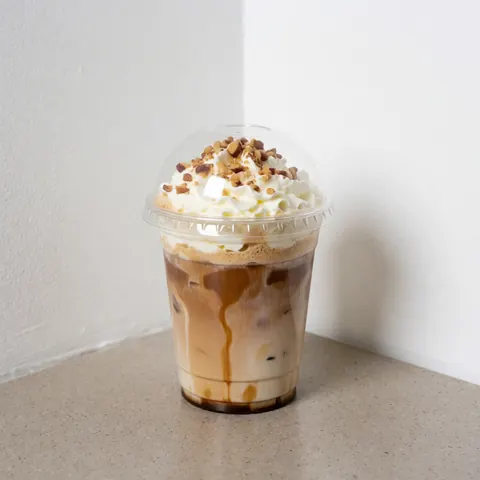 Iced coffee in a clear glass topped with whipped cream and chopped nuts on a light stone surface with white walls and natural lighting