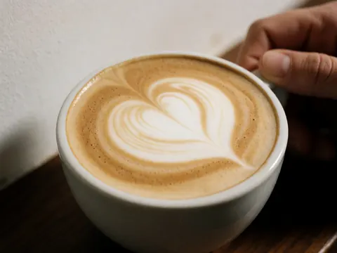 Cappuccino with heart latte art in a white cup held by hands on a dark wooden table against a white background