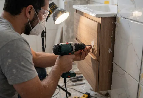 Carpenter installing a wooden bathroom vanity using an electric drill, safety gear, and work light in a remodeling bathroom.