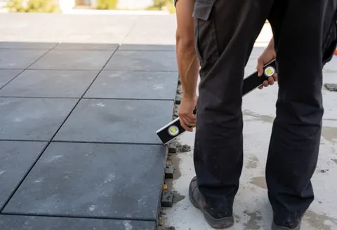 Worker using a level to align gray tiles on an outdoor terrace during daylight, ensuring precise installation.