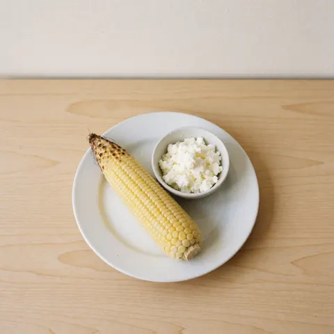 Cooked Peruvian corn topped with white cheese served on a white plate placed on a light wooden table with natural lighting