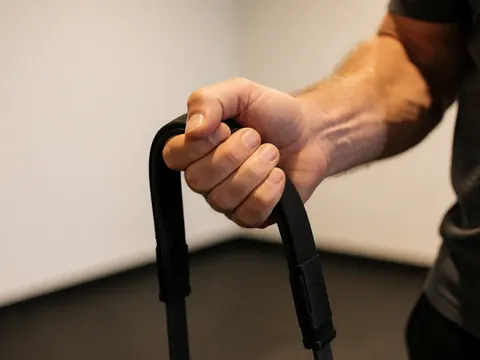 Detailed view of an adult hand holding a black resistance band, showing skin texture and material details against a white wall and gym floor background.
