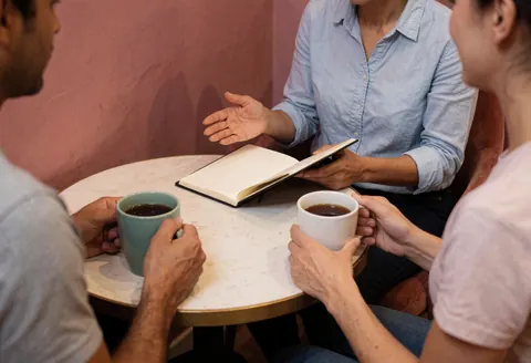 Personal trainer explains fitness plan to client at a small round table with coffee mugs and an open notebook in a warm setting