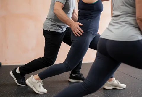 CrossFit coach correcting client's lunge posture in a gym with terracotta walls and rubber flooring under bright natural light