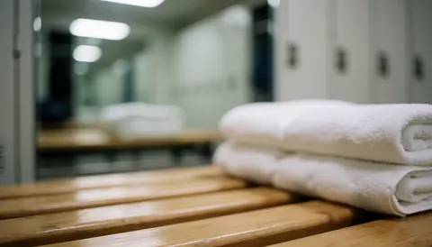 Folded white towels resting on a wooden bench in a clean gym locker room with blurred mirrors and lockers in the background
