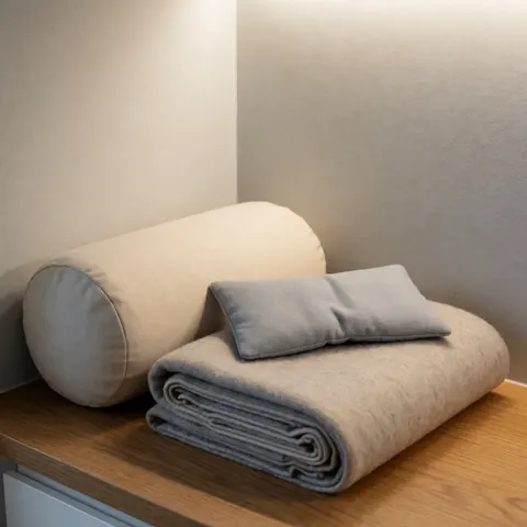 Restorative yoga kit featuring a cushion, blanket, and bolster pillow arranged on a wooden table against gray walls with warm lighting and natural textures.