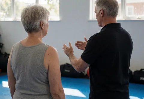 Back view of boxing instructor gesturing with hands while teaching footwork technique to an older female client in a gym with natural light and blue floor