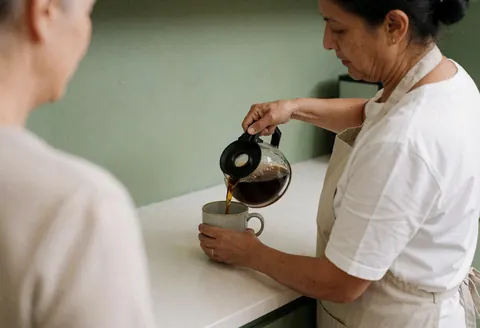 Coffee being poured from a glass carafe into a ceramic mug on a white table with a pale green wall in the background.