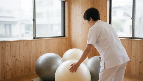 A physiotherapist in a white uniform adjusts three stability balls in a studio with wooden walls and natural light.