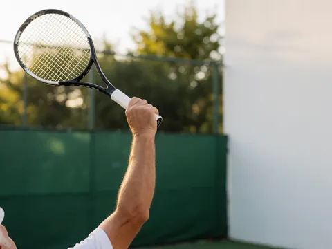 Extended arm holding a tennis racket in motion during a serve, with a green fence and white wall background under warm afternoon light