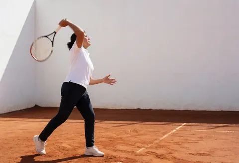 A female tennis player in action hitting a smash on a red clay court with a white wall background
