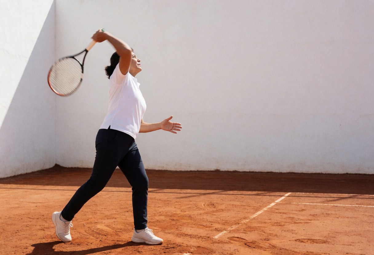 A female tennis player in action hitting a smash on a red clay court with a white wall background