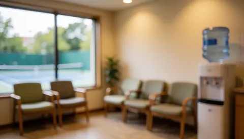 A comfortable waiting space featuring upholstered chairs, a water dispenser, and a window overlooking a tennis court with warm lighting and a relaxed atmosphere.
