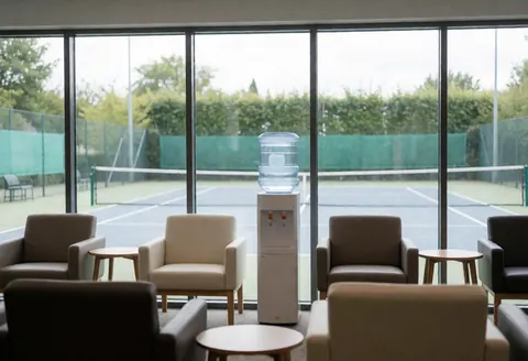 Modern waiting room with comfortable chairs, side tables, and a water dispenser overlooking an outdoor tennis court through large windows.