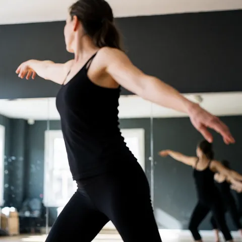 A dancer in black clothing performs a dynamic pose in a studio with gray walls and reflective mirrors.