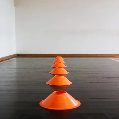 Orange training cones lined up on a dark wooden floor with white walls, ideal for agility exercises and basketball drills