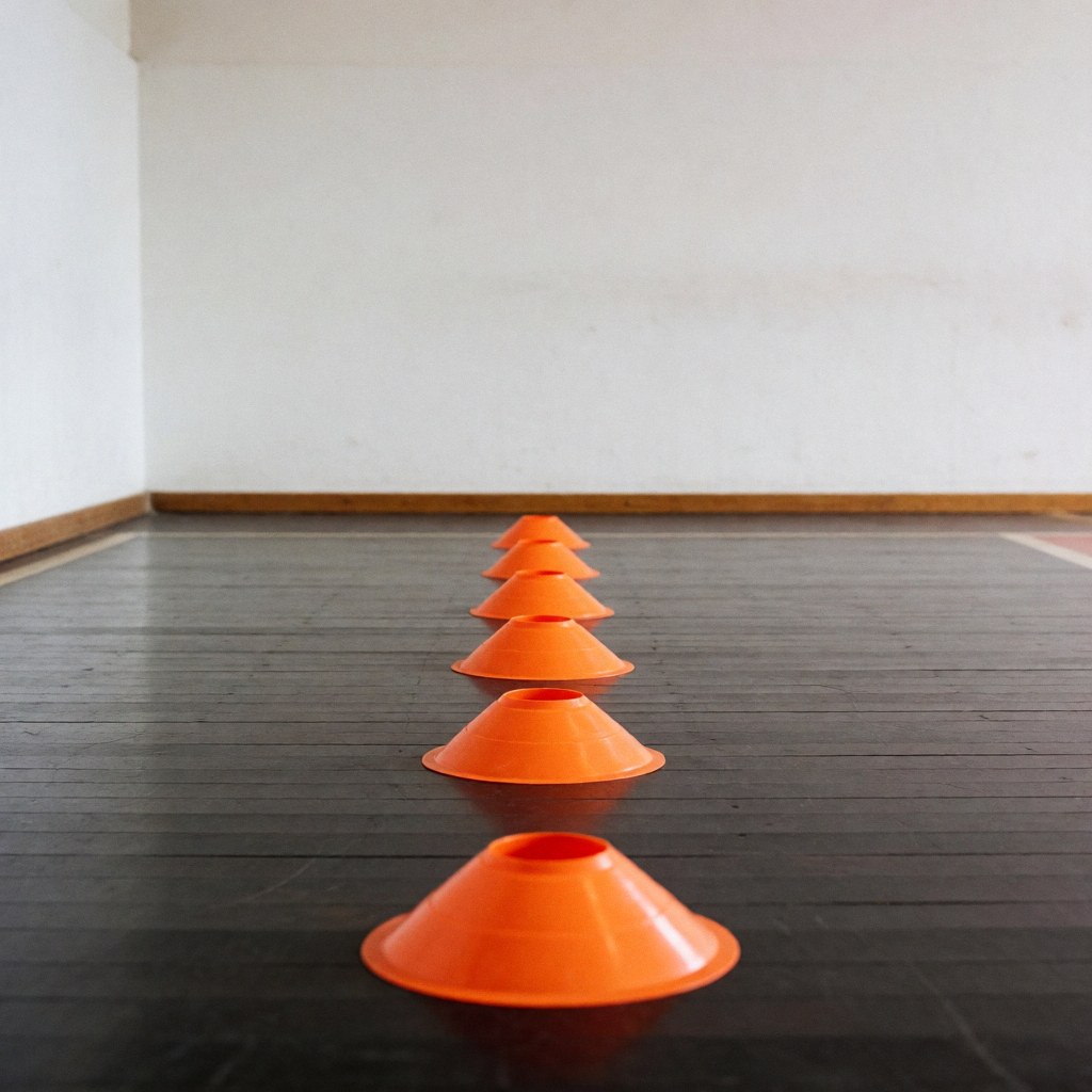 Orange training cones lined up on a dark wooden floor with white walls, ideal for agility exercises and basketball drills
