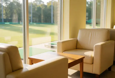 Beige armchairs and wooden table in a golf waiting room with light yellow walls and natural light overlooking the course