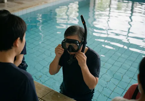Swimming coach adjusting a mask and snorkel tube by the poolside while students watch from the side.