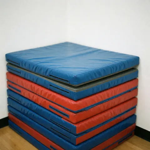 Stack of red and blue gymnastics mats in a gym corner with wooden floor and white wall, bright lighting and visible texture.