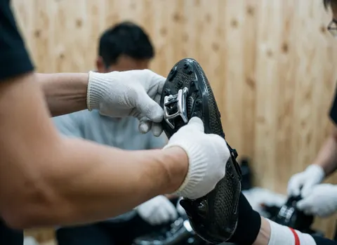 Close-up of gloved hands tightening a carbon cleat on a cycling shoe with wooden walls in the background