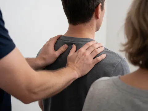 A therapist applies a shoulder massage to a client while another person observes in a bright neutral setting