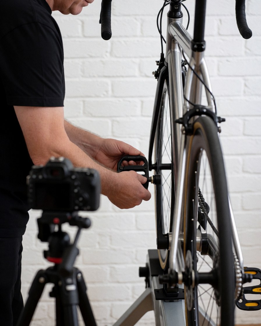 Close-up of a technician adjusting a bicycle pedal on a repair stand against a white brick wall with a camera on a tripod nearby