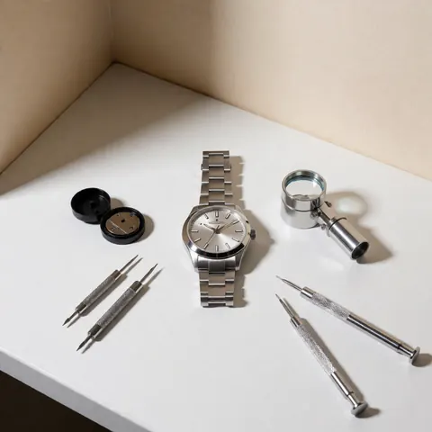 Silver wristwatch accompanied by repair tools and a magnifying glass on a white surface, suitable for watchmaking catalogs.