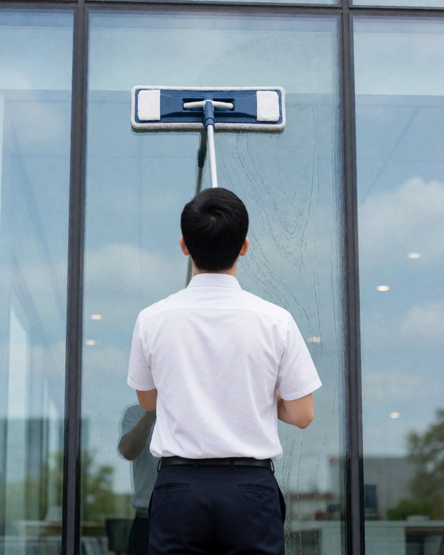 Worker in white shirt and dark pants cleaning an office window with a squeegee and microfiber mop against a modern glass building.
