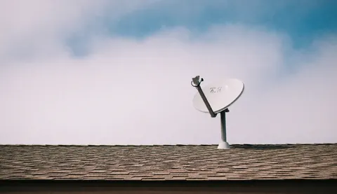 White satellite dish mounted on a shingled roof against a blue and cloudy sky, suitable for antenna installation service backgrounds.