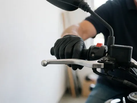 Close-up of a hand wearing a black glove adjusting a motorcycle brake lever in a workshop with blurred background and visible tools