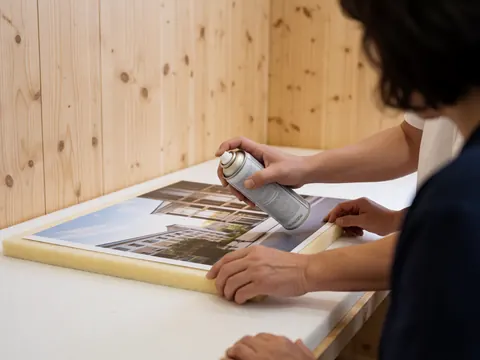 Close-up of a hand applying spray adhesive to a printed graphic on a foam board in a workshop setting
